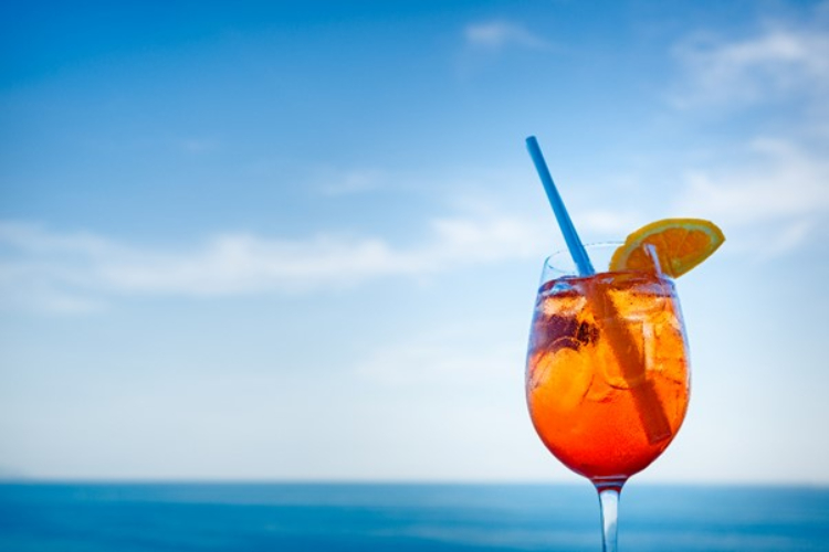 A glass of Aperol Spritz with a straw with the ocean on the background